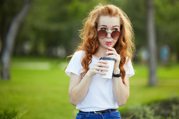 beautiful red-haired girl walks through the park with a cup of coffee. The girl is smiling at the camera. A girl in sunglasses and a modern watch on her arm