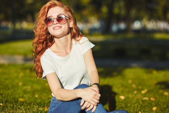 A lovely red-haired girl, warmed by the rays of the sun, is sitting on a lawn and posing for the camera. The girl is wearing a T-shirt with jeans, glasses on her face, and a modern gadget on her arm