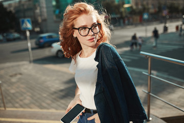 Beautiful red-haired girl stands in the street on the background of the city and posing on camera....