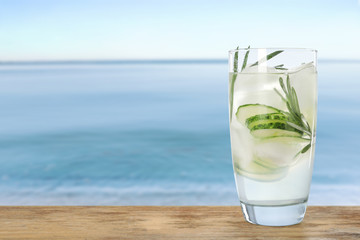 Glass of refreshing drink with cucumber slices and rosemary on wooden table near sea, space for text