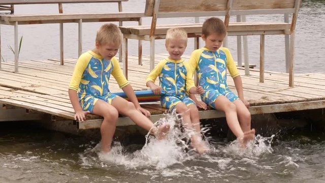 Funny children in bathing suits sit together on wooden bridge, legs in water. boys are happy on holiday in village together. Summer day, river, swimming in water. Closeup of legs and feet