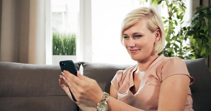 Glamorous lady sitting on couch and scrolling smartphone, looking photos in new application