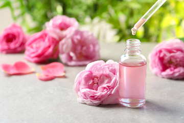 Dripping rose essential oil into bottle with pipette and fresh flowers on grey table, space for text