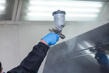 A male worker in jumpsuit and blue gloves paints with a spray gun a side part of the car body in black after being damaged at an accident. Auto service industry professions