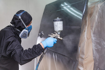 A male worker in jumpsuit and blue gloves paints with a spray gun a side part of the car body in...