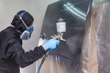 A male worker in jumpsuit and blue gloves paints with a spray gun a side part of the car body in black after being damaged at an accident. Auto service industry professions