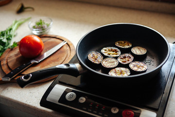 Eggplants are fried in a skillet. Vegetables on a cutting board in the background.