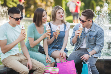 Group of young cheerful people eating ice cream and having fun. Shoppers outdoors. People after shopping. Funny group of friends resting near the fountain.