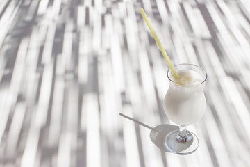 Pina colada cocktail with yellow drinking straw, coconut milk and ice cubes on white bar table. Sunlight and shadow play. Alcohol drink in glass. Summer vacation, refreshment concept.