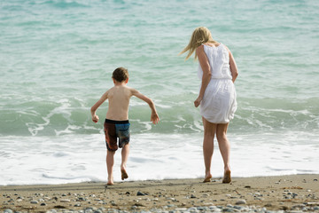 Fototapeta na wymiar Blonde slim mother and her child enjoy together summer, waves of sea at beach.