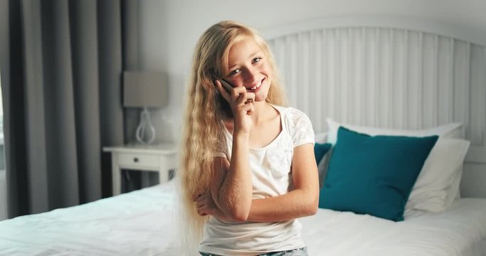 Beautiful long girl dressed in white t-shirt sitting on bed and talking on smartphone, smiling and feeling happy