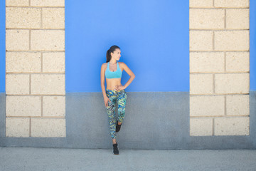 Full body fit Caucasian woman wearing a blue sports bra and leaning against a wall, posing with a pensive, relaxed look after an intense workout routine, healthy lifestyle, sport or fitness concept