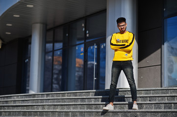 Asian man in eyewear and yellow sweatshirt posing against new modern building.