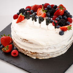 Closeup of a festive homemade cake on the table. Layered cream vanilla cake decorated with berries of raspberry, strawberry and blueberry. Candles the word HAPPY in the foreground. Celebration concept