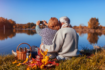 Senior couple taking selfie while having picnic by autumn lake. Happy man and woman enjoying nature and hugging