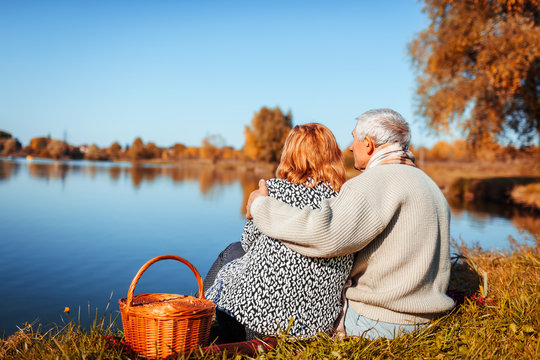 Senior couple having picnic by autumn lake. Happy man and woman enjoying nature and hugging