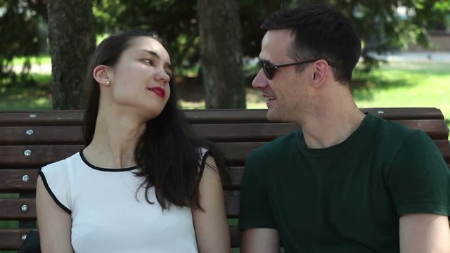 A young couple of lovers are talking and laughing while sitting on a bench in the shade of trees. A young attractive girl in a white T-shirt and a young handsome guy in sunglasses and a green T-shirt