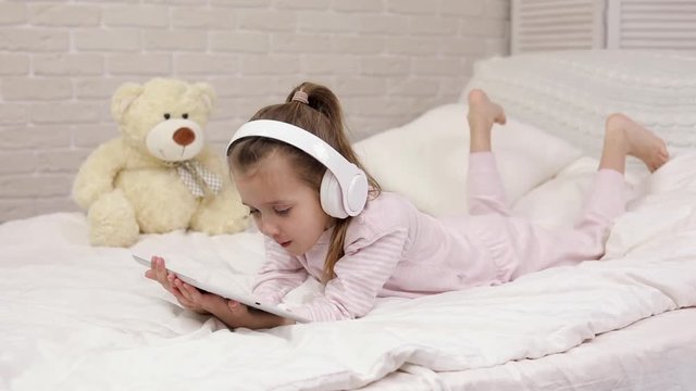 little child baby girl listening to the music with the headphones and digital tablet on bed.