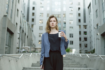 Fototapeta premium Pondering busy caucasian businesswoman looking aside while holding coffee in urban setting. Thoughtful office worker holds laptop and coffee cup near the business center concrete stairs, looking away.