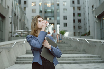 Attractive young office worker drinking coffee looking up holding the laptop. Thoughtful businesswoman drinks coffee near the office building standing on the stairs with the computer.