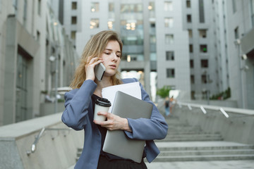 Young confident business woman walking in hurry downstairs, holding documents, laptop, coffee cup, talking on mobile phone on city street in front of modern office building. Horizontal, copy space.