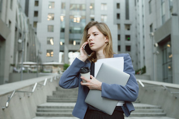 Busy caucasian woman having phone business discussion while holding coffee cup, papers and laptop near office building. Office worker standing on stairs with smartphone, computer, documents and coffee