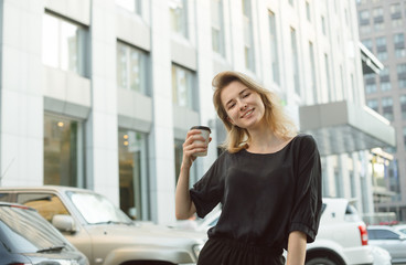 Smiling happy young woman holding a cup of coffee, wearing grey top and black trousers. Youthful cheerful girl in front of the urban scenery and car parking. Broad smile. - Powered by Adobe