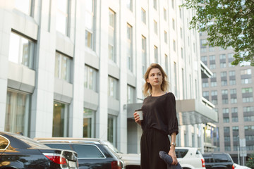 Portrait of calm elegant woman looking aside dressed in grey blouse and plack pants holding coffee in front of skyscraper. Young stylish girl walking down the street in downtown with take away coffee.
