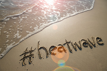 Modern message for the beach weather, heatwave, with a social media-friendly hashtag written in smooth sand with lens flare over an incoming wave