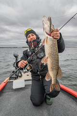 Pike fishing. Happy fisherman with big fish trophy at the boat with tackles