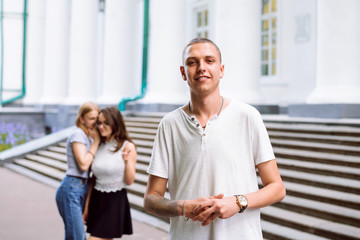 Fototapeta premium Handsome student standing in front of college building while girls are gossiping about him, flirts and staring at him