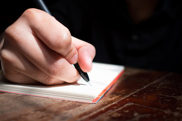 Hand is writing a book on a wooden table near a window. Close up.