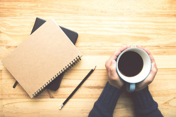 A woman wearing a blue shirt holding a coffee cup with two hands. And there are two books, a pencil on a wooden table near the window.