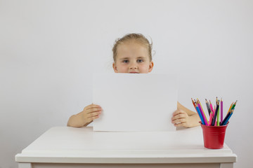 Little schoolgirl sitting at her desk t holding a white sheet of paper, space for text