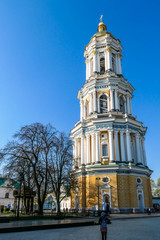 Fototapeta na wymiar A girl in a beanie standing in front of a four storied bell tower of Pechersk Lavra complex in Kiev, Ukraine. The tower is golden an white. Fe trees growing around it. Clear blue sky.