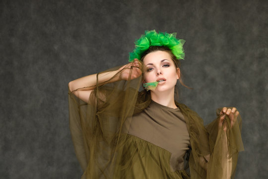 Portrait in lower chest on a gray background of a pretty young brunette woman with a green floral wreath in her hair. Standing in different poses, talking, showing hands, demonstrating emotions.