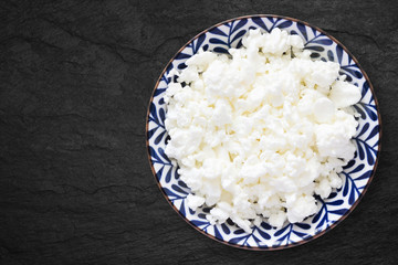 An overhead photo of fresh natural cottage cheese in a floral ceramic bowl on the black stone desk. Organic eco healthy meal, dairy product. Top view. Place for text, copyspase.