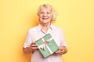 beautiful old lady holding a gift box and smiling. isolated yellow background. studio shot....