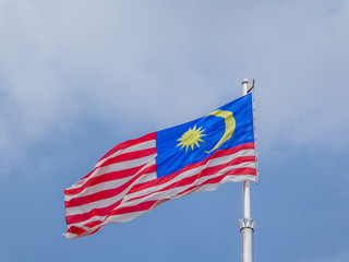 A Malaysian flag waving proudly on the mast. Flag consists of red and white stripes, blue square with golden moon and sun. Flag contrasts with cloudy sky in the back.