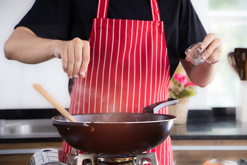 A man cooking food in the kitchen and adding some seasoning into a cooking pan