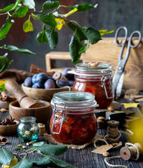Two glass jars with homemade canned plums jam, marmalade, jelly on rustic wooden table with cardamon, cinnamon,  anise, bowl of plums