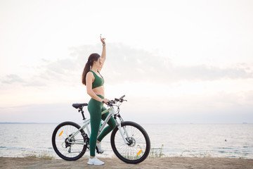 Fototapeta premium Beautiful young brunette woman resting after bicycle ride at sunrise. Model listening to music with wireless earbuds in the morning.