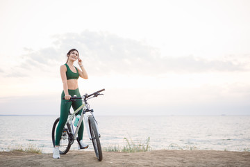 Obraz premium Beautiful young brunette woman resting after bicycle ride at sunrise. Model listening to music with wireless earbuds in the morning.