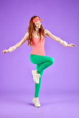 red-headed girl with freckles in pink bodysuit, green leggins and pink sun cap doing aerobics exercises keeping one leg raised and looks down with smile, enjoys training. 80s style aerobics concept