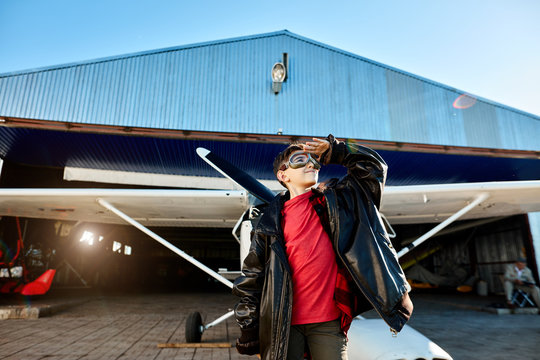 Cute Little Kid Stands Outside Hangar Building Near Single-engine Airplane, Waiting His Dad To Land At Airfield, Dressed In Father's Pilot Jacket And Aviator Glasses. Aviation And Family Concept.