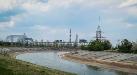 Fototapeta premium A remote view on the infamous Unit 4 Reactor in Chernobyl, Ukraine. Lush green grass growing in front of the reactor. Clean river flowing in the nearby. Reactor is covered with sarcophagus. Overcast
