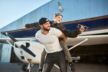 happy little boy in aviator glasses sitting on father's back, playing with toy airplane, enjoying sunny summer day and time together, light propeller plane and hangar on the background. © alfa27