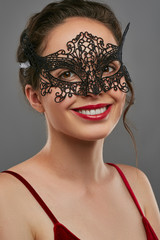 Half-turn shot of smiling lady with dark hair, wearing wine red crop top. The woman is tilting her head and looking at camera, wearing black carnival mask with perforation, tied with satin ribbon.