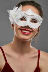 Portrait of smiling lady with tied back dark hair, wearing wine red crop top. The girl is raising her head, wearing snow-white carnival mask, adorned with beads and massive flower made of feathers.