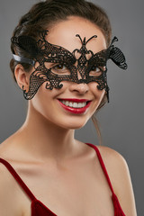 Obraz premium Half-turn shot of woman with tied back dark hair, wearing wine red crop top. The smiling lady is tilting her head, wearing black masquerade mask with fancy perforation. Vintage carnival accessory.